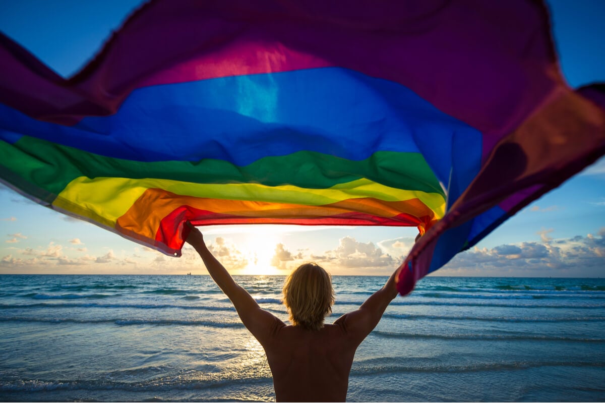 Ein Mann mit einer Regenbogenflagge am Strand feiert seine LGB T-Reise.